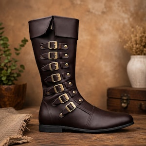 May include: A brown leather knee-high boot with multiple gold-colored buckles and a fold-over top. The boot has a pointed toe and is set against a neutral background. The boot is approximately 16 inches tall.
