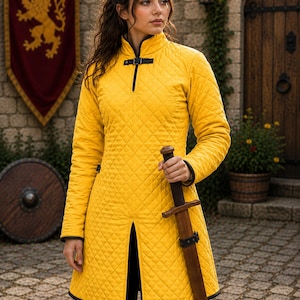 May include: A bright yellow quilted tunic featuring a high collar, black trim, and a black buckle closure at the neckline. The garment has long sleeves and a front slit, and is paired with a wooden sword held in a vertical position.