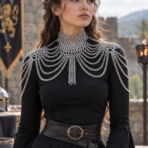 May include: A black long-sleeved dress with flared sleeves and a high neckline, adorned with a silver-toned chainmail shoulder piece featuring multiple draped chain layers. A wide, dark brown leather belt with a circular metal buckle is worn.