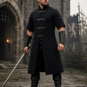 May include: A black quilted long tunic featuring a high collar, short sleeves, and four front buckle closures. The garment is paired with black leather bracers on the forearms and black leather boots. A long steel sword is held in the right hand.