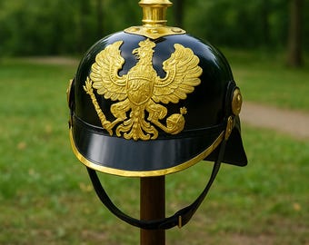 May include: A black and gold military helmet with a prominent gold eagle emblem. The helmet features a pointed top and a chin strap. It is displayed on a wooden stand against a blurred green background.