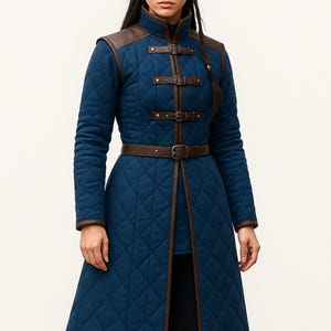 Könnte beinhalten: Ein langer, gesteppter blauer Mantel mit braunen Lederdetails. Der Mantel hat einen hohen Kragen, Schulterpartien und einen Gürtel. Das Design umfasst Rautensteppung und dekorative Schnallen. Ein stilvolles Oberbekleidungsstück.