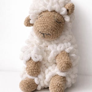 May include: A crocheted stuffed sheep toy featuring a soft, white, looped yarn body and head covering. The face, ears, arms, and legs are crafted from a smooth, light brown yarn. The sheep has embroidered closed eyes and a small mouth.