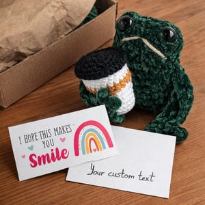 May include: A handmade green crochet frog holding a crochet coffee cup with a black lid and orange stripe. A card with a rainbow and the text "I hope this makes you smile" is next to a blank card. The items are on a wooden surface.