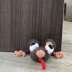 May include: A crocheted dark gray creature with a flat, wide body, featuring two large white eyes with black pupils, a light pink nose, and a bright red tongue sticking out. The creature has small light pink hands extending from its sides.