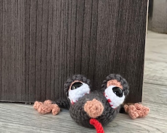 May include: A crocheted dark gray creature with a flat, wide body, featuring two large white eyes with black pupils, a light pink nose, and a bright red tongue sticking out. The creature has small light pink hands extending from its sides.