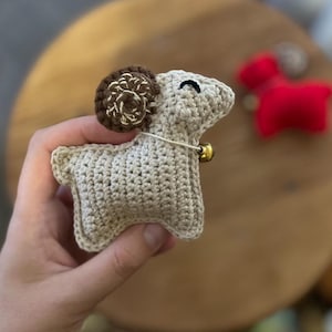 Könnte beinhalten: Eine kleine, beige gehäkelte Widder-Figur mit einem dunkelbraunen, spiralförmigen Horn und einem einzelnen schwarzen, gestickten Auge. Eine dünne Schnur ist um den Hals gebunden und hält eine kleine, goldfarbene Metallglocke am Körper.