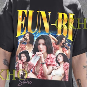 May include: Black Unisex T-Shirt with a graphic of a woman in a red and white checkered top. The shirt features the name "EUN-BI" in gold lettering with a lightning bolt design. The shirt also has the text "OKHO Store".