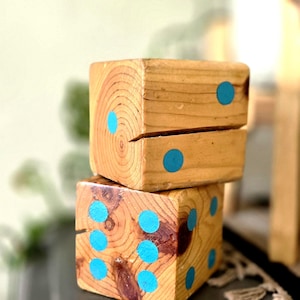 May include: Two wooden dice stacked on top of each other. Each die is a natural wood color with visible grain and blue dots. The dice are sitting on a dark gray surface.