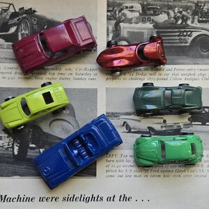 May include: Six toy cars in various colors, including red, green, blue, and yellow, are arranged on a newspaper page. The cars are positioned near black and white images of vintage race cars. The newspaper text is partially visible.