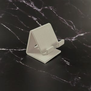 Könnte beinhalten: Ein hellgrauer Telefonständer mit geripptem Design. Der Ständer ist abgewinkelt und hat eine Ablage zur Aufnahme eines Telefons. Der Ständer besteht aus einem festen Material und befindet sich auf einer schwarz-weißen Marmoroberfläche.