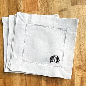 May include: Three white linen cocktail napkins with a decorative stitched border. Each napkin features a black embroidered image of a dog's head in the corner. The napkins are stacked on a wooden surface.