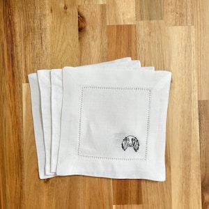 May include: Four white linen cocktail napkins, each with a square embroidered border and a black embroidered dog's head. The napkins are stacked on a wooden surface.