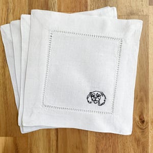 Könnte beinhalten: Stapel von vier weißen quadratischen Cocktailservietten aus Leinen. Jede Serviette hat eine dekorative Naht und ein kleines gesticktes schwarz-weißes Hunde-Gesicht. Ideal für Getränke.