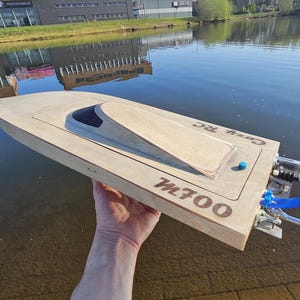 Op de afbeelding: Een lichtgekleurde houten op afstand bestuurbare boot met de tekst M700 en Cozy RC gegraveerd op het dek. Het vaartuig heeft een donkere cockpitkap, een kleine blauwe knop en een metalen propellerconstructie met felblauwe slangen aan de achterkant.