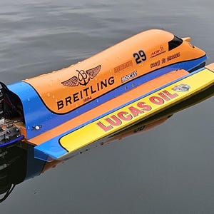 Op de afbeelding: Een model speedboot op het water, met oranje, blauwe en gele details. De boot toont het nummer 29, de tekst "BREITLING 1884" en "LUCAS OIL". Het is een gedetailleerd model van een raceboot.