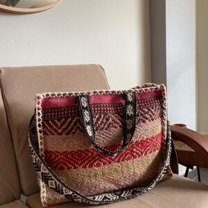 May include: A woven tote bag with a geometric pattern in shades of red, pink, beige, and brown. The bag has a long, patterned strap and a matching woven design on the top edge. The bag is sitting on a beige cushion.