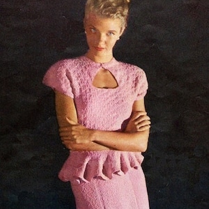 May include: A woman wearing a pink knitted dress with a keyhole neckline and a ruffled skirt. She is standing in front of a dark background and holding her arms crossed. A blue and green bowl is on a wooden table in front of her.