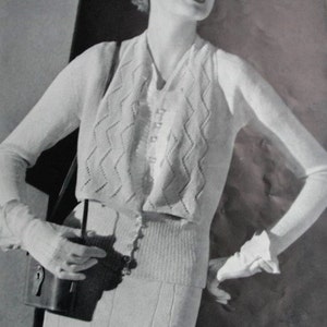 May include: A black and white photo of a woman wearing a knitted cardigan and skirt set. The cardigan has a chevron pattern and the skirt has vertical ribs. The woman is wearing a hat and holding a small purse.