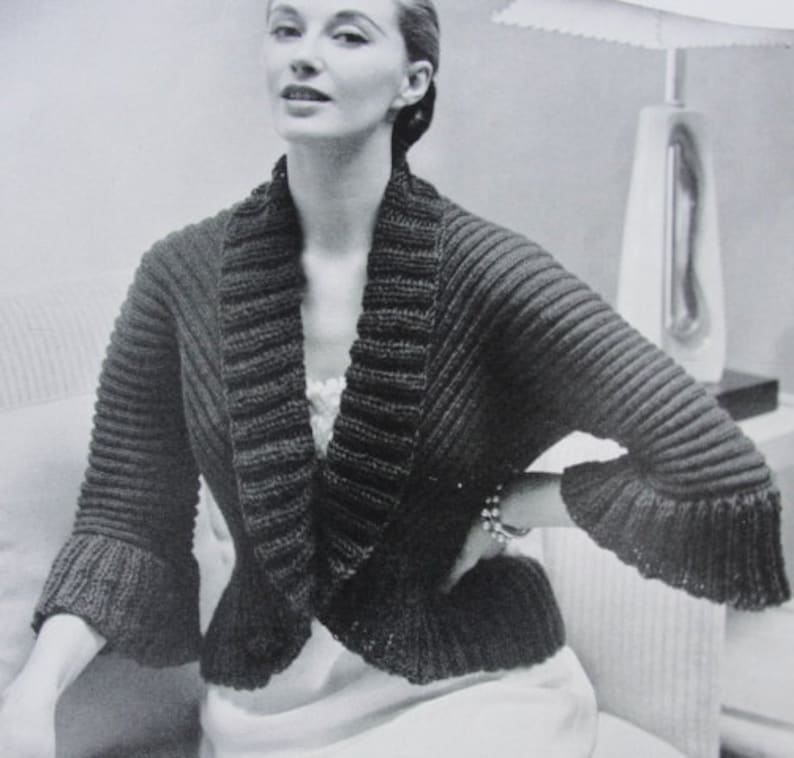 May include: A black and white image of a knitted bolero jacket with a wide collar and flared sleeves. The jacket has a ribbed texture and is worn by a person. The bolero is a vintage style.
