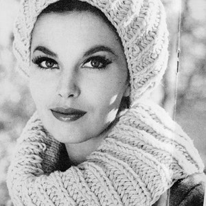 May include: A black and white photo of a woman wearing a white knitted hat and matching scarf. The hat is a slouchy beanie style and the scarf is a thick, chunky knit.