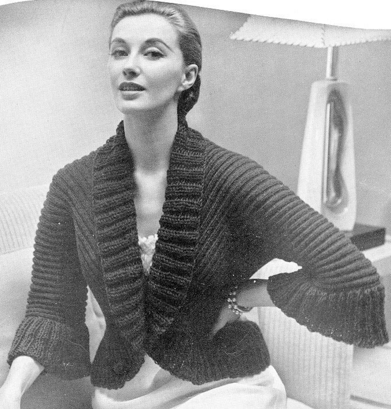 May include: A dark brown, ribbed-knit bolero jacket with a wide collar and flared sleeves. The jacket is worn over a white garment. The sleeves have a ruffled edge. The jacket is a vintage style.