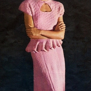 May include: A woman wearing a pink knitted dress with a keyhole neckline and a ruffled skirt. She is standing in front of a dark background and holding her arms crossed. A blue and gold bowl is on a table in front of her.