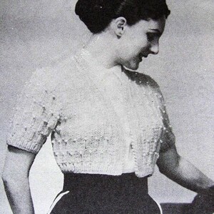 Può includere: Un cardigan bianco lavorato a maglia con maniche corte e chiusura con bottoni. Il cardigan è indossato sopra una gonna nera.