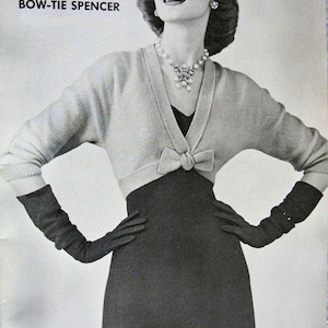 May include: A black and white photo of a woman wearing a light gray knitted cardigan with a bow tie detail. The cardigan is cropped and worn over a black dress. The woman is wearing a pearl necklace and earrings. The text "BOW-TIE SPENCER" is printed at the top of the image.