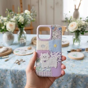 May include: A patchwork phone case with a lavender border and a white terrier applique. The case features various fabric patterns, including floral and solid colours, and a small yellow star. The case is held in a hand.