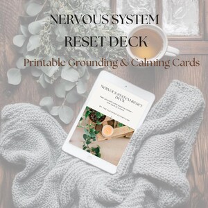 May include: A digital tablet showing "NERVOUS SYSTEM RESET DECK" and "Printable Grounding & Calming Cards." The tablet is on a grey knitted blanket, with a cup of tea and foliage in the background.