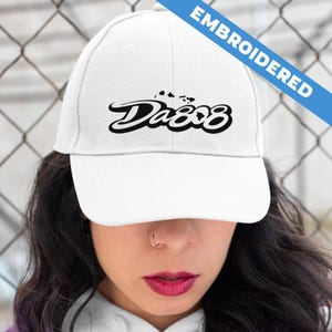 May include: A white baseball cap featuring black embroidered text that reads Da808 with a small graphic of the Hawaiian Islands above the lettering. A blue diagonal banner in the upper right corner contains the white text EMBROIDERED.
