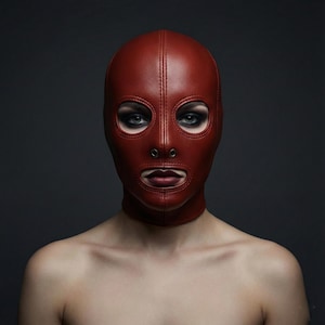 May include: A red leather hood with eye and mouth openings. The hood has a vertical seam down the centre and a neck strap. The person is wearing dark eye makeup.
