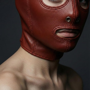 May include: A close-up of a red leather hood with eye and mouth openings. The hood is stitched and has a neck collar. The background is a dark grey.