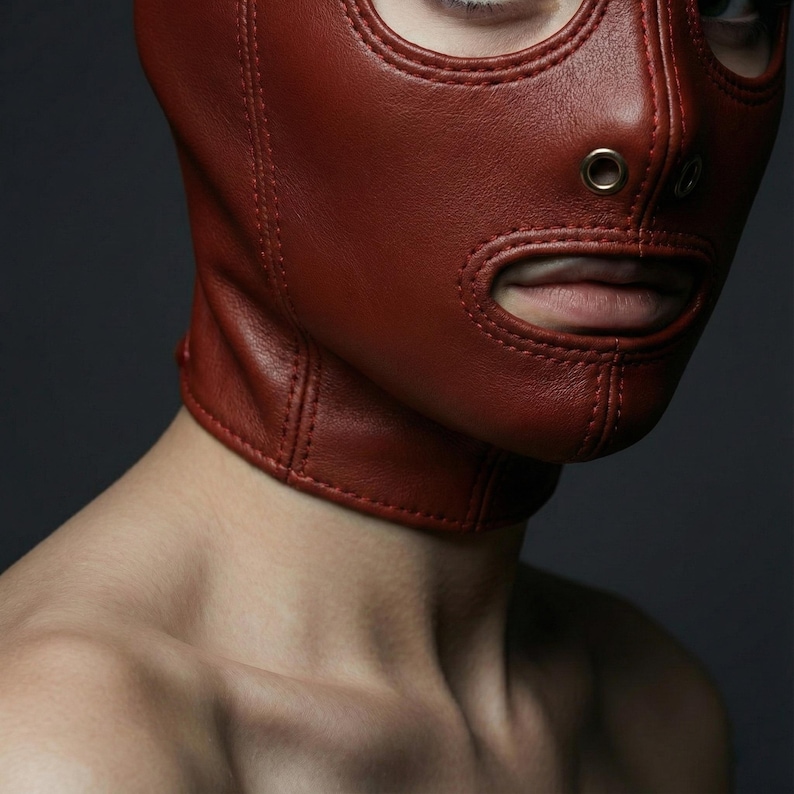 May include: A close-up of a red leather hood with eye and mouth openings. The hood is stitched and has a neck collar. The background is a dark grey.