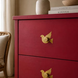 May include: A deep red wooden chest of drawers featuring two drawers, each fitted with a matte gold bird-shaped metal handle. The bird handles are positioned centrally on the drawer fronts, showcasing a silhouette of a bird in flight with a circular knob.