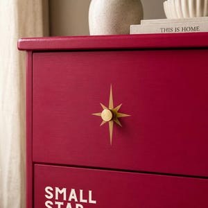 May include: A magenta-coloured chest of drawers featuring a gold-toned starburst-shaped drawer pull. The lower drawer displays the white text "SMALL STAR" in a sans-serif font. The surface is smooth with a matte finish and clean, straight edges.