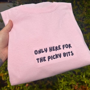 Op de afbeelding: Een lichtroze crewneck T-shirt met de donkerblauwe geborduurde tekst "ONLY HERE FOR THE PICKY BITS". Het gevouwen shirt wordt vastgehouden, met groene bladeren op de achtergrond.