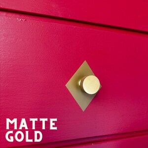 Op de afbeelding: Een close-up van een felroze kastdeur met een matte gouden knop. De knop is rond met een geribbelde rand, gemonteerd op een ruitvormige plaat. De woorden "MATTE GOLD" zijn in het wit gedrukt.