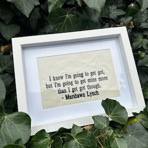May include: A white framed print featuring the quote "I know I'm going to get got, but I'm going to get mine more than I get got though. - Marshawn Lynch" on a beige background. The frame is rectangular.
