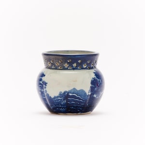 Puede incluir: Un jarrón de cerámica azul y blanco con un diseño de paisaje. El jarrón tiene un borde azul oscuro con recortes decorativos y un interior azul claro. El cuerpo del jarrón presenta una detallada escena de paisaje azul y blanco.