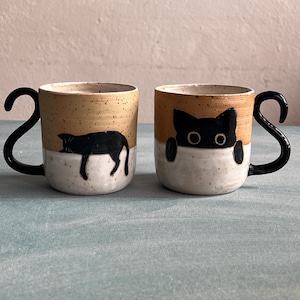 Könnte beinhalten: Zwei Keramikbecher mit schwarzen Katzenmotiven. Die Becher haben eine gesprenkelte beige obere Hälfte und eine weiße untere Hälfte. Jeder Becher hat einen schwarzen Henkel. Ein Becher zeigt eine schlafende Katze, der andere eine Katze, die über den Rand schaut.