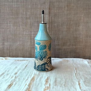 May include: A ceramic oil bottle with a light blue top and a stainless steel spout. The body of the bottle is beige with a teal leaf pattern. The bottle is sitting on a white cloth.