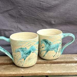 May include: Two ceramic mugs with a cream base and turquoise horse designs. The mugs have turquoise handles and a textured, artistic appearance. The mugs are placed on a wooden surface.