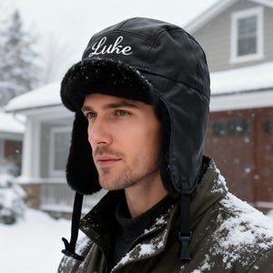 May include: Black winter hat with ear flaps and a faux fur lining. The word "Luke" is embroidered in white script on the front of the hat. The hat has a buckle closure under the chin.