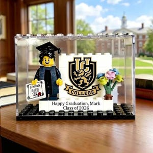 May include: A Lego graduation display featuring a minifigure in a cap and gown holding a certificate. The display includes a white sign with a college crest and the text "Happy Graduation, Mark Class of 2026". A small bouquet of flowers is also included.