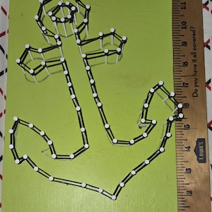 May include: A handmade anchor string art piece on a green background. The anchor is outlined with black string and white nails. The artwork is approximately 10 inches tall and 7 inches wide, perfect for nautical decor.