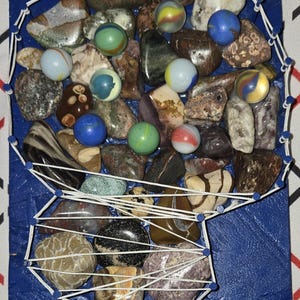 Puede incluir: Un tablero rectangular azul con un diseño de arte con hilos que perfila la forma de una cabeza humana. El interior de la cabeza está lleno de una colección de varias piedras de río lisas y multicolores y varias canicas de vidrio.