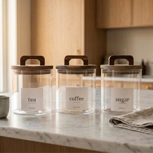 May include: Three clear glass cylindrical canisters with wooden lids featuring rectangular handles. Each canister has a white label with black text reading tea, coffee, or sugar. A small grey ceramic cup and a folded beige linen cloth are nearby.
