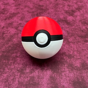 Puede incluir: Una Poké Ball roja, blanca y negra, un dispositivo esférico utilizado en la franquicia Pokémon para capturar y contener Pokémon. La mitad superior es roja, la mitad inferior es blanca, y una banda negra y un botón circular detallan el centro.